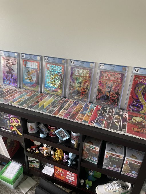 A dark wood display shelf showcasing graded comic books, including multiple issues of 'Defenders of the Earth,' 'Ghost Rider,' and 'TMNT,' along with various figurines and pop culture merchandise.