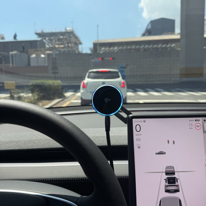 A view from inside a Tesla showing the steering wheel and central touchscreen displaying "0 km/h" and a "60" speed limit, with a Qi2 wireless charger mounted on the dashboard. The driver is stopped behind a white car at a red light, humorously complaining that Full Self-Driving (FSD) still isn't working despite the system apparently recognizing the speed limit.