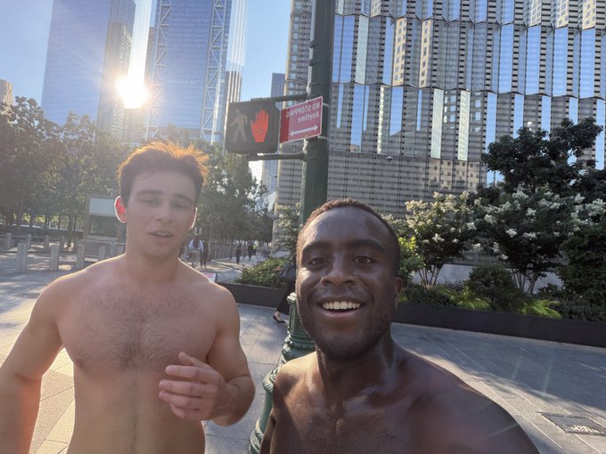 A selfie of two shirtless men standing on a city street with tall skyscrapers in the background, next to a pedestrian crossing signal showing a red hand ('Don't Walk').