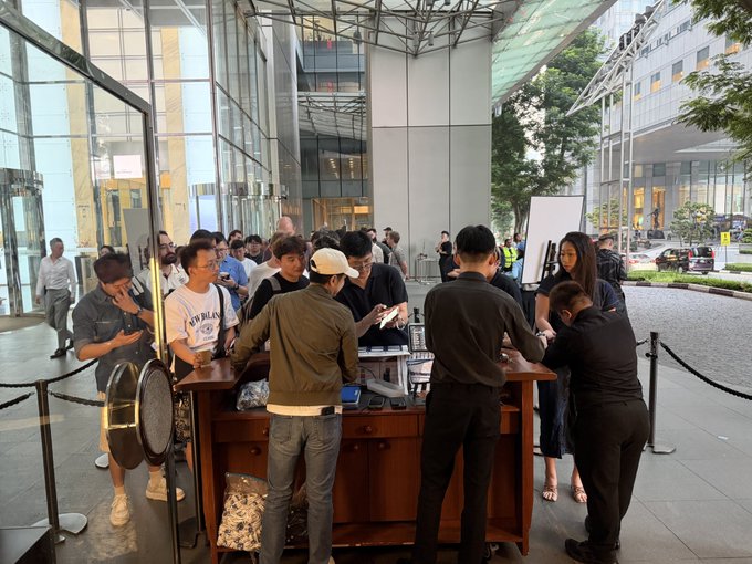 A crowd of people gathers around a wooden desk set up outside a modern glass building, where staff in black assist with a check-in or registration process.