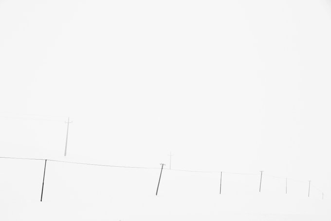 A hazy, minimalist landscape showing faint utility poles stretching across a misty, pale gray sky.