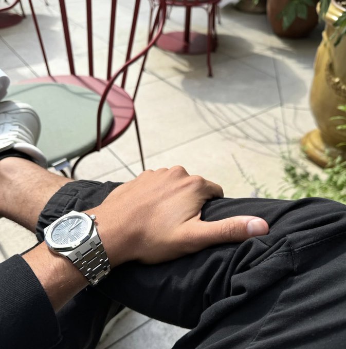 A close-up of a man's wrist wearing a silver watch while resting on his black pants at an outdoor café, paired with a post celebrating Solana ($SOL) hitting 0.75 and asking followers to like and say gm.