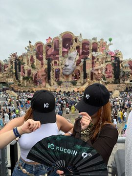 Two friends pose with a KuCoin-branded fan at the Tomorrowland festival, where a massive, ornate stage featuring giant sculpted faces towers over the crowd in the background, celebrating the brand's collaboration with the event.