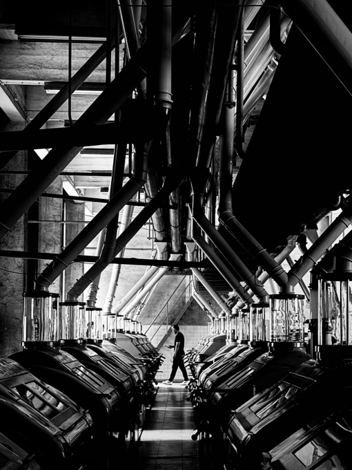 A black-and-white photograph shows a lone figure walking through an industrial corridor lined with massive pipes and machinery, creating dramatic symmetrical composition.