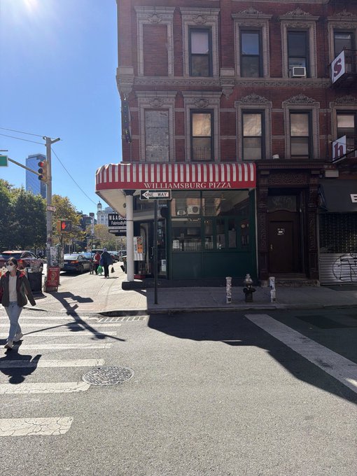 A street corner view of Williamsburg Pizza, featuring a red and white striped awning on a brick building under a clear blue sky.