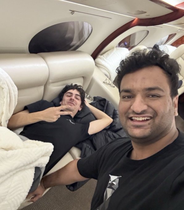 Two men take a selfie inside a private jet; one smiles in the foreground while the other lies down on a leather seat in the background making a peace sign.
