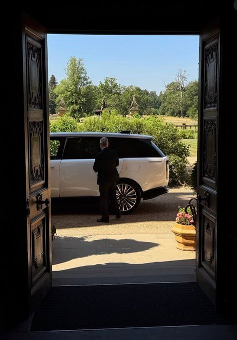 A view through an open ornate doorway shows a man in a dark suit standing outside next to a white Range Rover parked on a gravel driveway near a garden.