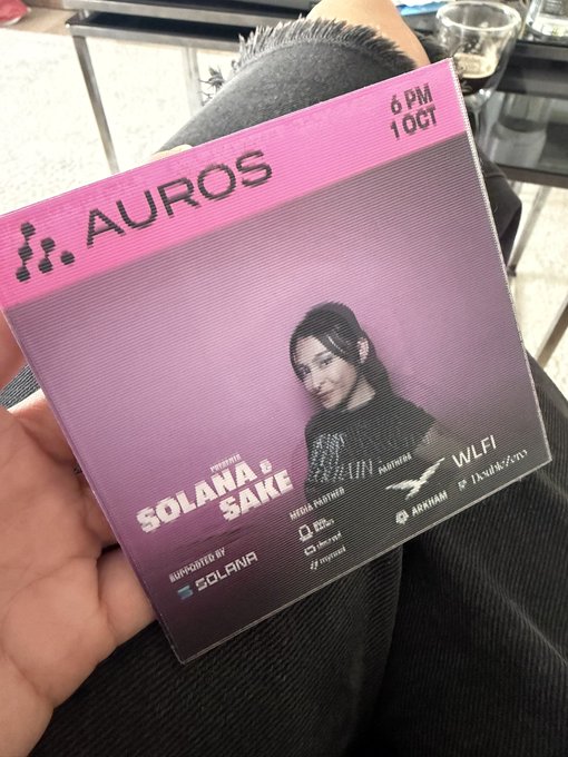 The image shows a person holding up a pink CD cover with a picture of a woman on it, along with some text that reads "Auros" and "Solaana Sake". The background appears to be a table or countertop. There is also a cup visible in the scene.