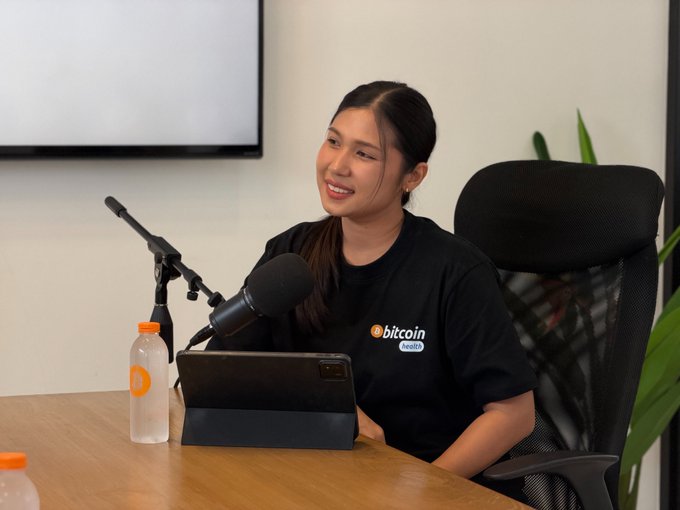 A smiling young woman wearing a black Bitcoin t-shirt sits at a desk with a microphone, tablet, and water bottle, appearing to record a podcast or interview.