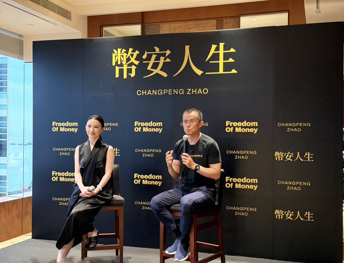 Two people sit on stools at a "Freedom Of Money" book talk event, with the backdrop displaying "幣安人生 CHANGPENG ZHAO" — Changpeng Zhao (CZ), co-founder of Binance, speaking into a microphone while sharing insights about his minimalist lifestyle and principles from his autobiography, as referenced in the accompanying post.
