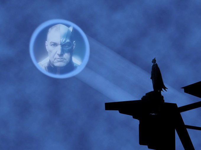A silhouette of a caped figure standing on a platform facing a glowing blue hologram of a man's head against a cloudy sky.