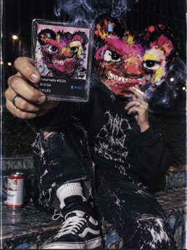 A stylized, edgy figure wearing a colorful mask and dark clothing holds up a card, sitting in a gritty urban environment with a spray can nearby.