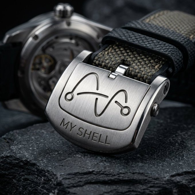A close-up of a wristwatch clasp featuring an engraved cat design and the text 'MY SHELL', attached to a canvas strap next to a skeleton-style watch face.