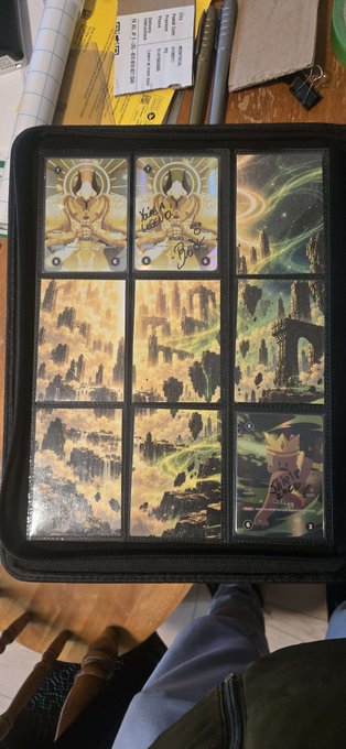 A 3x3 grid of illustrated cards depicting fantasy scenes and characters, signed by an artist, rests in a black zippered binder.