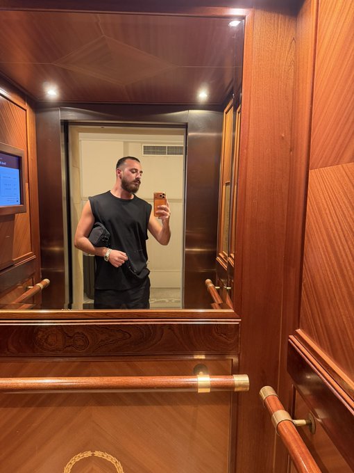 A man in a black sleeveless shirt takes a mirror selfie with his phone in an opulent elevator featuring rich wood paneling and brass handrails, checking his reflection as a stylish "vibe check." The luxury setting, complete with a small screen and elegant geometric wood patterns, perfectly complements the confident, fashion-forward mood of the moment.