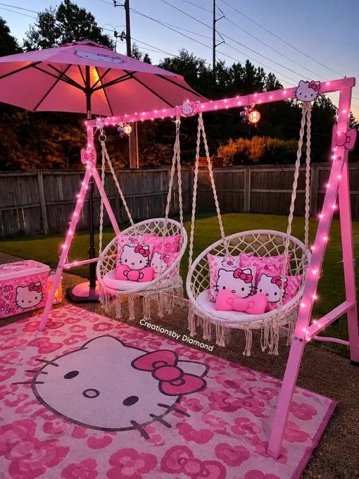 An outdoor Hello Kitty-themed patio setup featuring a pink swing set with hanging chairs, a matching umbrella, and a large Hello Kitty rug, all decorated with string lights and watermarked 'Creationsby_Diamond'.