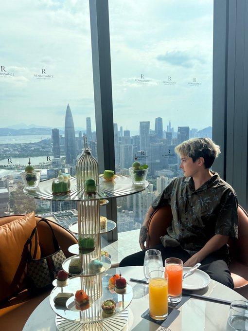 A person sits at a high-rise table with a tiered dessert stand and colorful drinks, overlooking a city skyline with the text 'RENAISSANCE' repeated on the window.