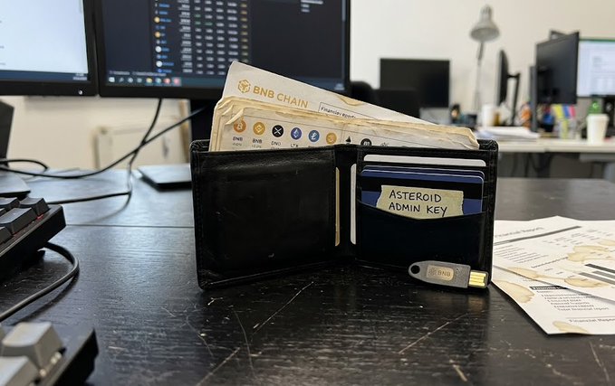 A photo of a black leather wallet open on a desk, containing cash and a 'BNB CHAIN' financial report, with a hardware wallet labeled 'BNB' and a card reading 'ASTEROID ADMIN KEY' tucked inside, alongside computer monitors in the background.