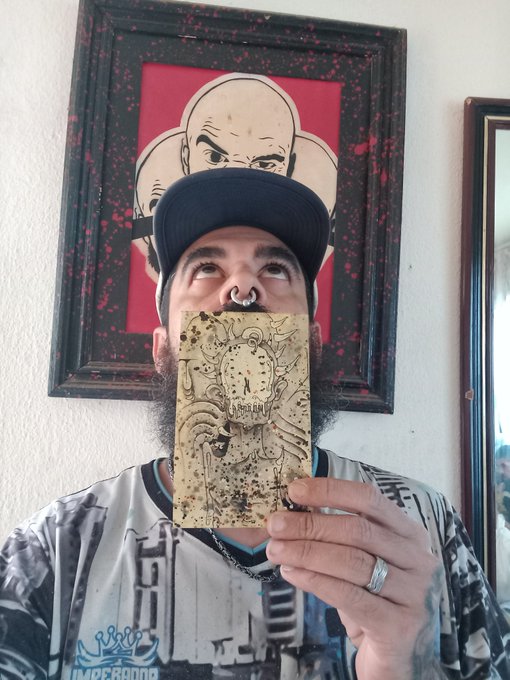 A bearded man wearing a cap covering a framed Mr. Bean artwork behind him, holding up a small horror-themed illustration that covers his mouth and nose, wearing an 'Imperioo' shirt.