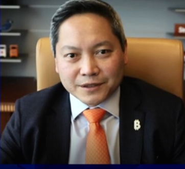 A man in a dark suit with an orange tie, wearing a 'B' lapel pin, sits in a tan leather chair.