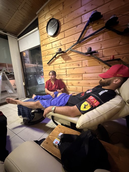 A man in a Ferrari racing jersey and cap relaxes barefoot on a couch while a woman sits cross-legged on the floor in a wood-paneled room, capturing a cozy, laid-back "vibe check" moment with scattered snacks and bags nearby.