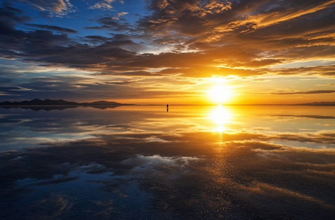 A scenic photograph of a sunset over a calm body of water (salt flat), with golden sunlight reflecting on the shallow wet surface and clouds streaking the sky.