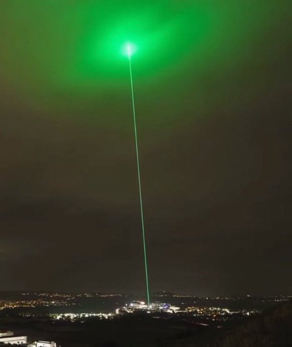 A bright green laser beam shoots from a city at night into the sky, illuminating the clouds with a vibrant glow. The image's "laser-to-the-moon" visual style echoes the post's promotion of a memecoin poised for massive 500x gains.