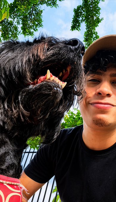 A young man wearing a tan cap and black t-shirt smiles for a selfie next to a fluffy black dog baring its teeth, matching the "With Nahla" caption as he poses with his playful pup outdoors.