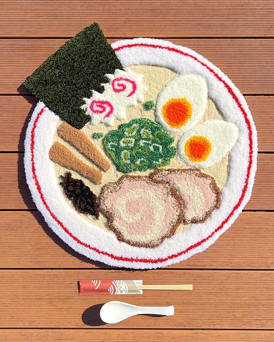 A creative rug designed to look like a bowl of ramen with nori, eggs, and meat sits on a wooden floor, accompanied by a matching chopstick holder and spoon rug.