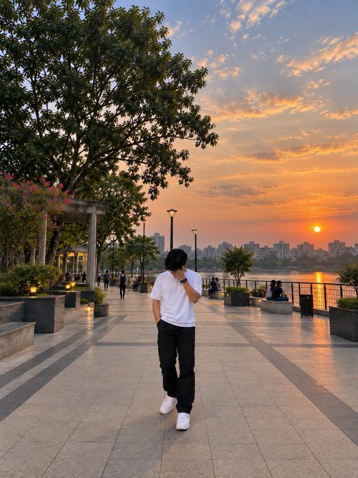 A person in a white t-shirt and black pants walking along a paved waterfront promenade at sunset, with trees, street lamps, and a city skyline across the water in the background.