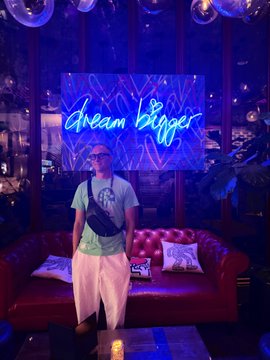 A person wearing a white cap, green t-shirt, and white shorts stands in front of a red leather couch beneath a neon sign reading "dream bigger" in blue and pink lighting. The post humorously introduces them as "me" with their "doginal dog," suggesting this is their first selfie rather than the intended dog photo.