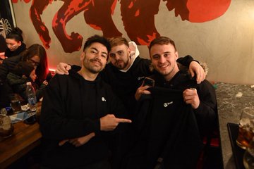 Three smiling men pose together at a viewing party, with one holding up a black "River" branded bag, celebrating an Argentina vs. Cape Verde match that ended 3-2 in extra time, hosted by RiverdotInc alongside River4fun and the $RIVER token.