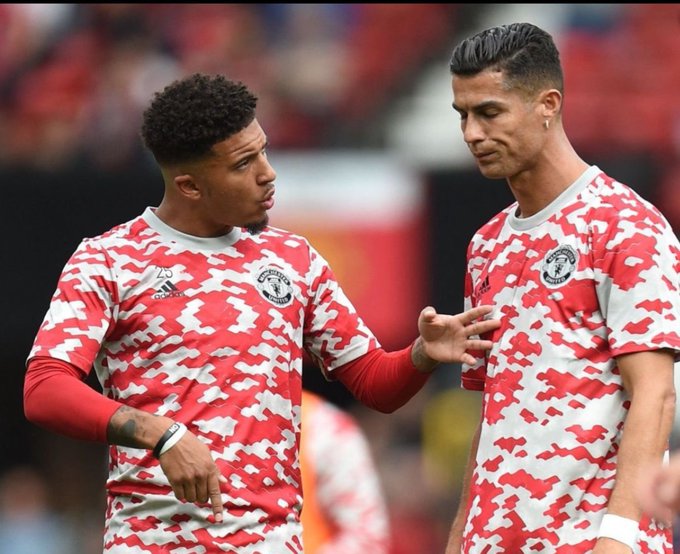 Manchester United players Jadon Sancho and Cristiano Ronaldo in red and white camouflage pre-match jerseys, with Sancho appearing to speak to Ronaldo.