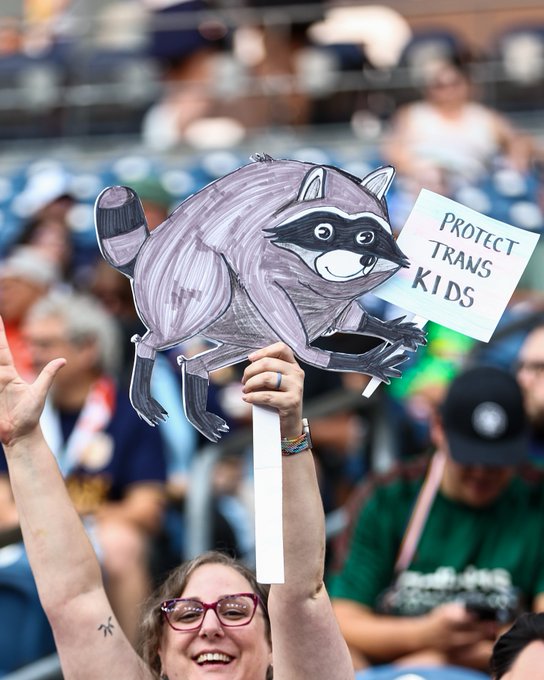 A woman smiling while holding up a hand-drawn cardboard sign depicting a raccoon holding a smaller sign that reads 'PROTECT TRANS KIDS,' with a crowd of people visible in the blurred background.