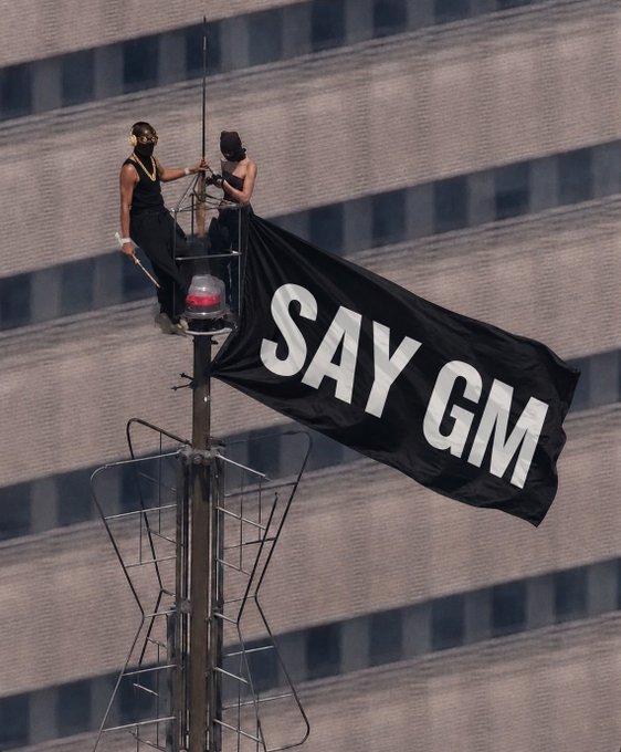 Two people sit atop a tall pole structure, unfurling a large black banner reading "SAY GM" against a backdrop of high-rise buildings.