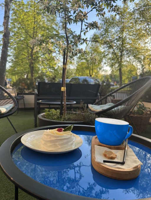 A slice of cake topped with whipped cream and a raspberry sits on a white plate next to a blue coffee cup on a wooden tray, placed on a blue table at an outdoor café setting.