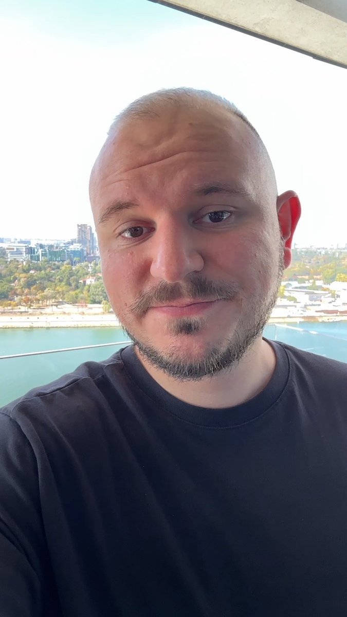 A close-up selfie of a bald man with a goatee wearing a black shirt, taken outdoors with a coastal city view in the background.