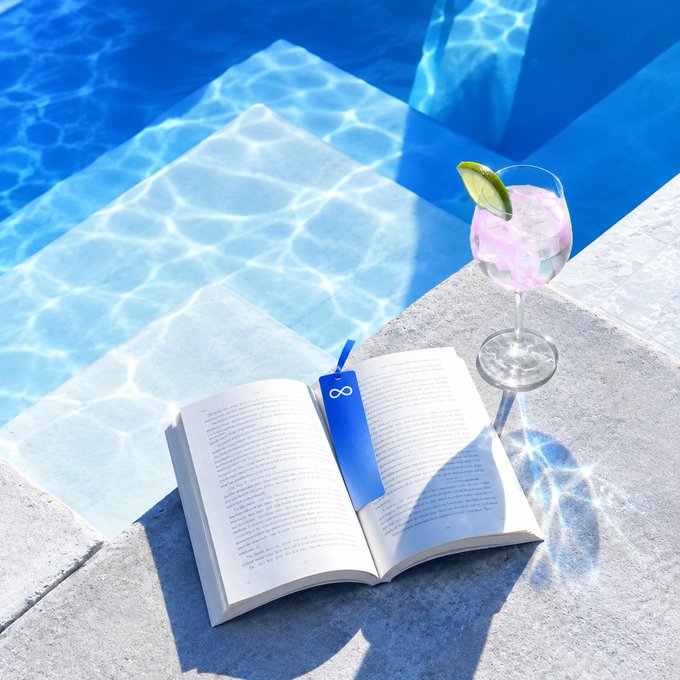 An open book with a blue bookmark marked "infinity/∞" rests on a poolside ledge beside a pink cocktail garnished with lime, capturing a relaxed moment of leisure by the water—perfectly matching the "Halfway in, No rush, GM" vibe of taking things slow.
