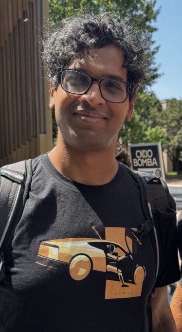 A smiling man with curly black hair and glasses wears a black t-shirt featuring a graphic of a car and the Tesla logo, with an 'OKO BOMBA' street sign behind him.