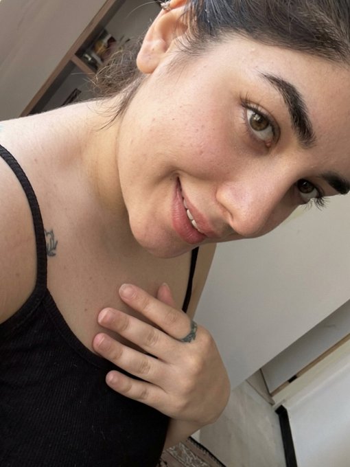 Close-up selfie photo of a young woman with dark hair pulled back, wearing a black top, with visible hand and shoulder tattoos.