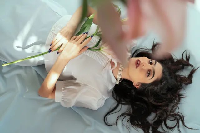 A woman with dark hair lies on a bed wearing a white blouse and pearl necklace, holding a green plant stem, with her face partially obscured by pink blurred elements.