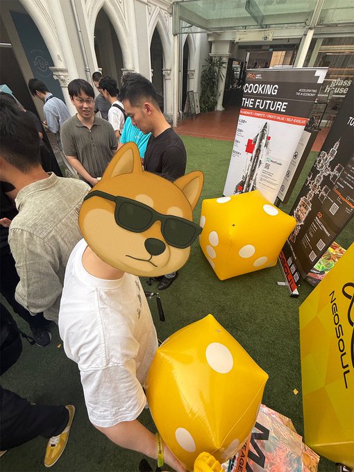 A person wearing a Shiba Inu dog mask with sunglasses at an indoor event with yellow dice props, with a banner visible reading 'COOKING THE FUTURE' and 'NASDUAL' branding.