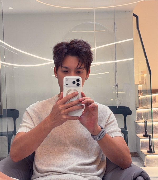 A man with styled brown hair takes a mirror selfie in a modern salon with a ring light, wearing a beige knit t-shirt and holding a white iPhone.