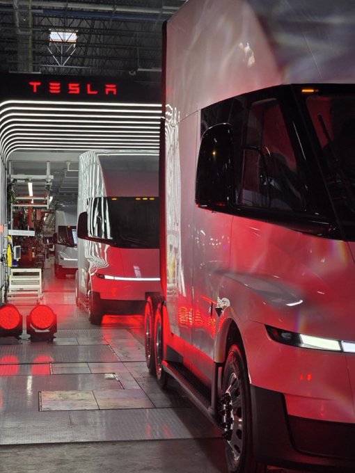 A row of red Tesla Semi trucks parked inside a manufacturing facility or loading area with the 'TESLA' logo visible on the wall.
