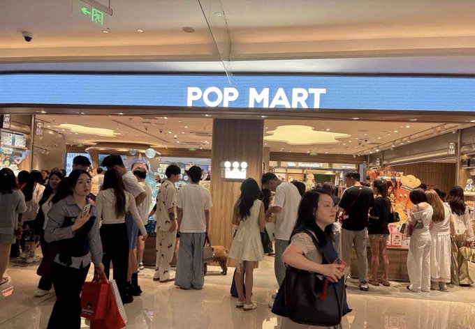 The image shows a large group of people gathered outside a store, possibly a pop art shop or a mall. The crowd consists of at least thirteen individuals standing close together, with some carrying handbags. A dog can also be seen among them. The scene suggests that there might have been an event or sale at the store, attracting many customers to visit and explore its offerings.