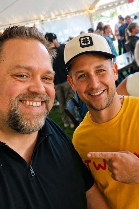Two smiling men pose for a selfie at what appears to be an outdoor event under a tent, with one wearing a white baseball cap featuring a logo and the other in a black shirt pointing at a yellow-shirted person.