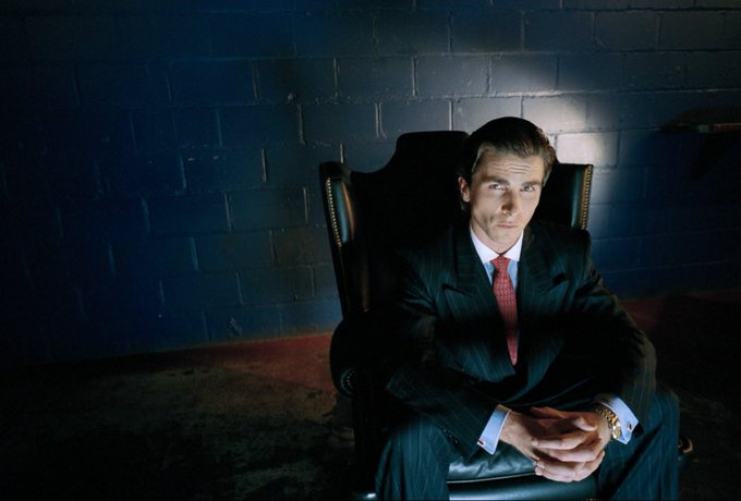 A man in a dark pinstripe suit with a red tie sits in a leather chair against a dark brick wall in a dramatically lit portrait.