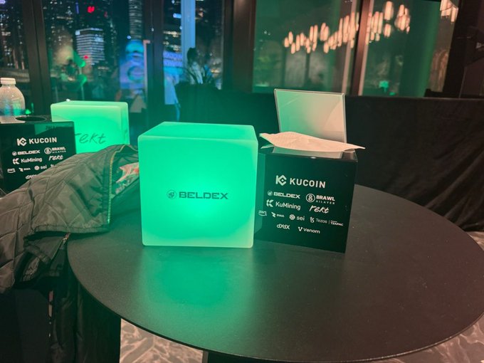 A glowing teal Beldex-branded cube sits on a round table beside a black Kuoin (KuCoin) sponsorship block featuring partner logos like Brawl, KuMining, and dYdX, likely from a crypto industry event. The illuminated cube highlights Beldex's branding, visually supporting the post's mention of $BDX's recent $8M funding and its push toward privacy infrastructure for Web3 and AI.