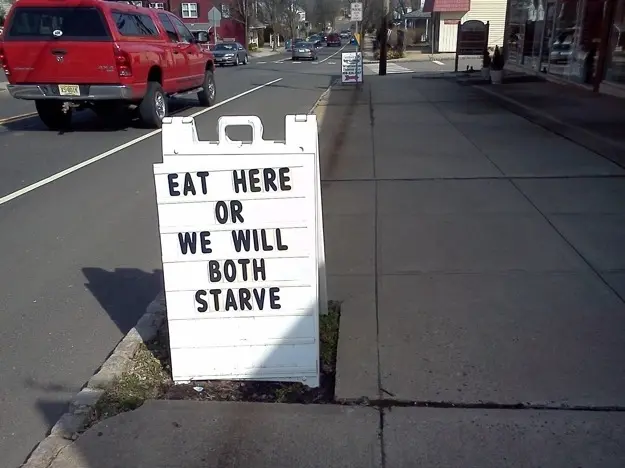 A white sandwich board sign on a sidewalk reading 'EAT HERE OR WE WILL BOTH STARVE' positioned along a street with a red pickup truck passing by.