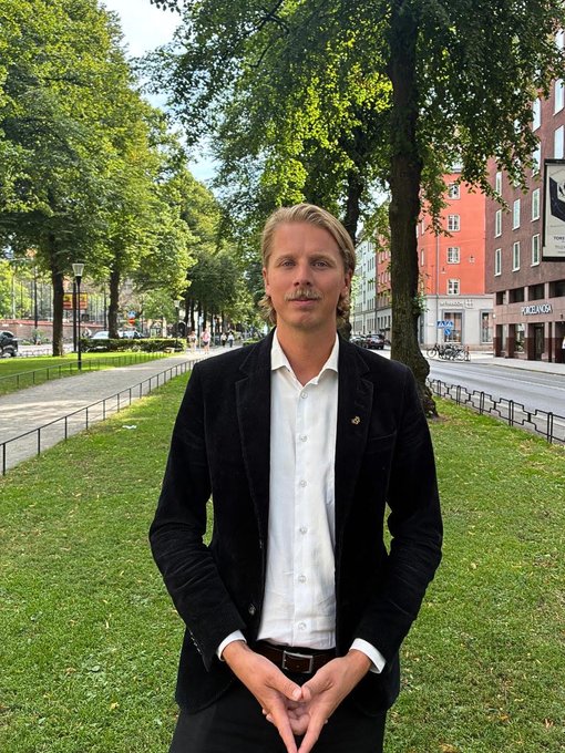 A blonde man in a black blazer and white shirt stands on a grassy urban pathway, with a "PRADA" store visible on a building behind him, complementing his announcement of stepping down as CEO of BTCX to become chairman of the board. The setting suggests a moment of reflection in the heart of a city, symbolizing his transition from hands-on leadership to a guiding role.